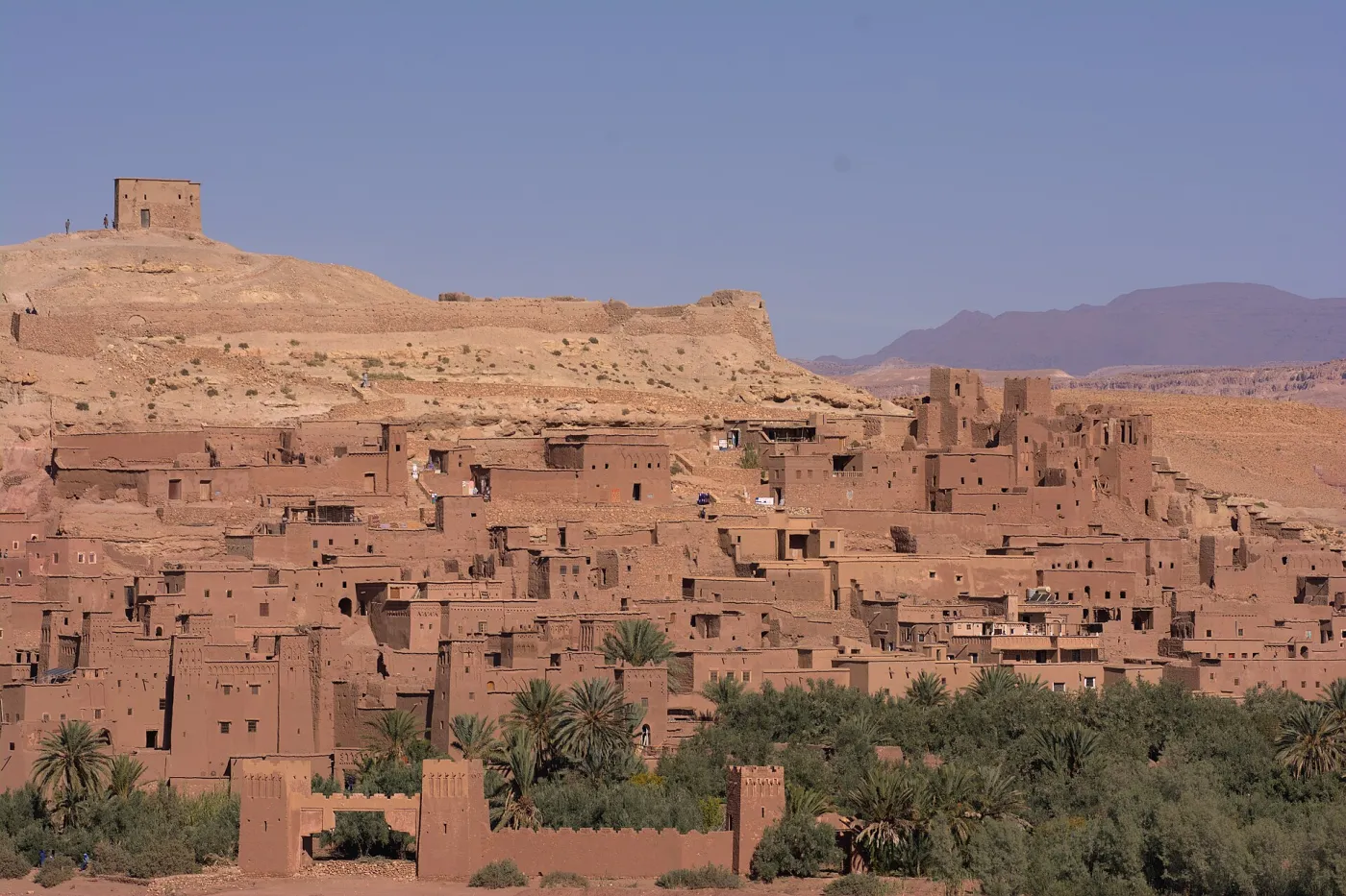 Earthen towers of the Ksar of Aït Benhaddou rising on a hill above the Ounila riverbed in golden late-afternoon light