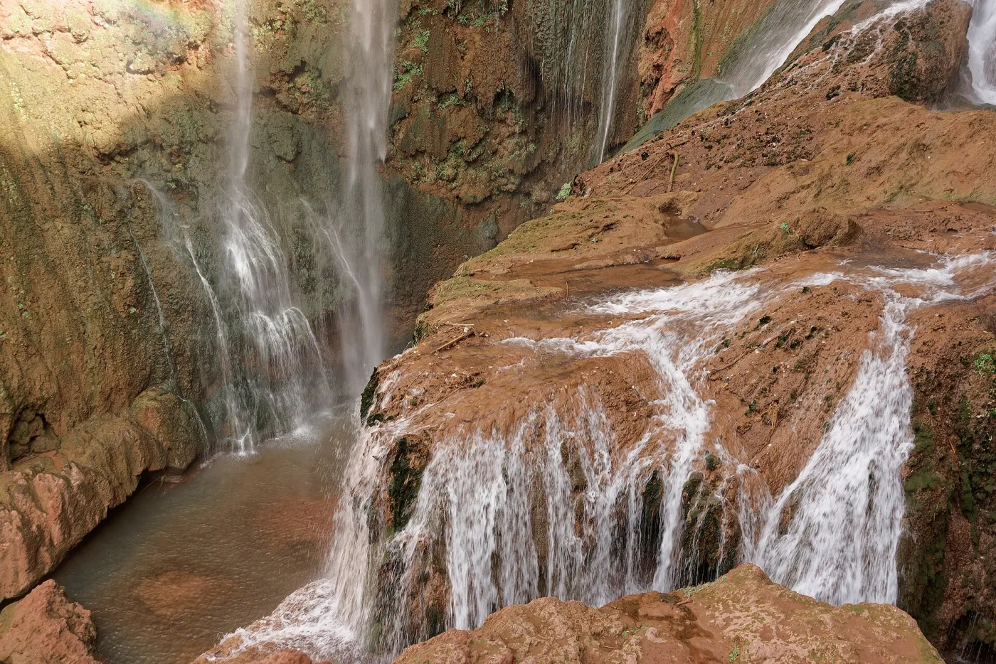 Tiered Ouzoud Waterfalls plunging into a green pool with small wooden rafts and a rainbow in the spray